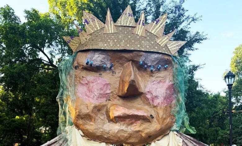 Giant handmade paper-mache figure with a spiky crown and pink cheeks outdoors in a park.