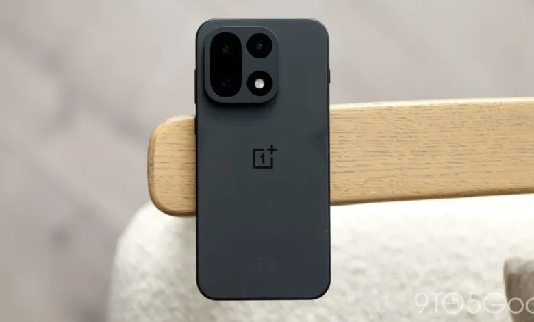 Black OnePlus smartphone resting upright against a wooden armrest in a bright setting.