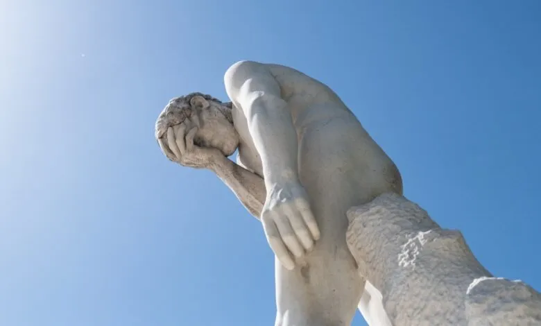 A classical white marble statue of a male figure holding his face in hand against a bright blue sky.