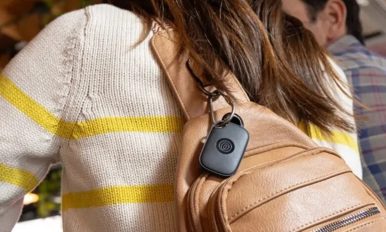 A black tracking device clipped to a tan sling backpack worn by a woman.
