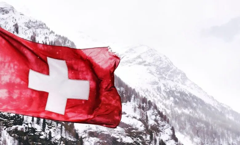 The Swiss flag waves proudly against a backdrop of majestic, snow-covered Alpine mountains.