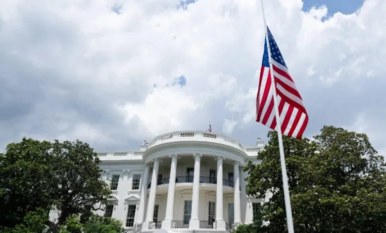The White House exterior with a large American flag flying against a cloudy sky.