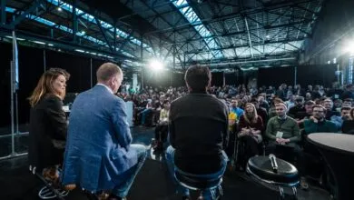 Panelists seated on stage speaking to a large audience in a large industrial warehouse venue.