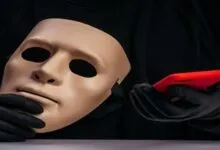 A person in a black sweater and gloves holds a blank mask and a red phone behind a keyboard.