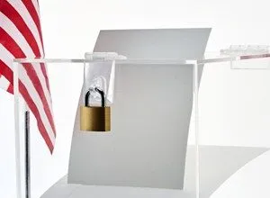 Clear acrylic ballot box secured with a brass padlock next to an American flag.