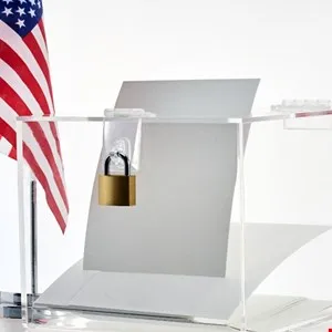 Clear acrylic ballot box secured with a brass padlock next to an American flag.