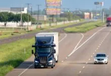 An autonomous semi-truck driving on a multi-lane highway next to a silver car.