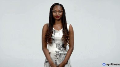 A Black woman with braided hair and glasses wearing a patterned dress stands against a light grey background.