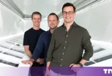Three male co-founders stand smiling in a modern, white, futuristic tech corridor.