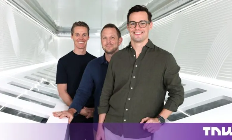 Three male co-founders stand smiling in a modern, white, futuristic tech corridor.
