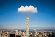 A lone fluffy white cloud hovers perfectly directly above a tall skyscraper in New York City.