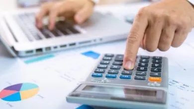 Close-up of a person using a calculator with financial charts and a laptop in the background.
