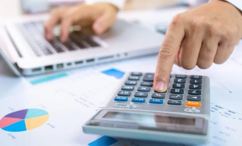 Close-up of a person using a calculator with financial charts and a laptop in the background.