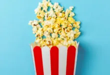 Red and white striped popcorn bucket overflowing with popcorn against a blue background.