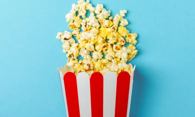 Red and white striped popcorn bucket overflowing with popcorn against a blue background.