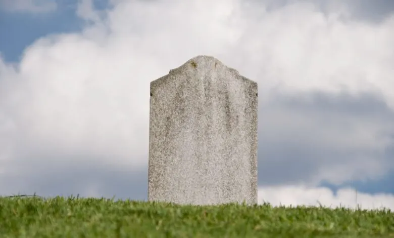 A blank, weathered white stone tombstone stands alone on a grassy hill against a cloudy blue sky.
