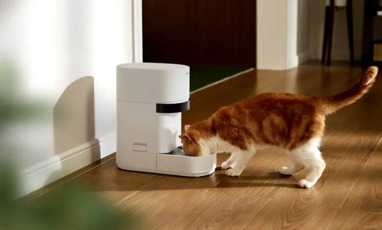An orange and white cat eats from a modern white automatic pet feeder on a wooden floor.