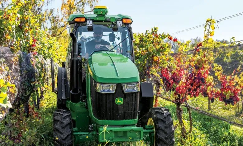 A John Deere 5ML tractor navigating rows of lush green and autumn-tinted grapevines in a vineyard.