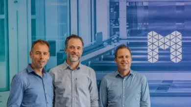Three men in button-down shirts stand smiling in front of a blue industrial background.