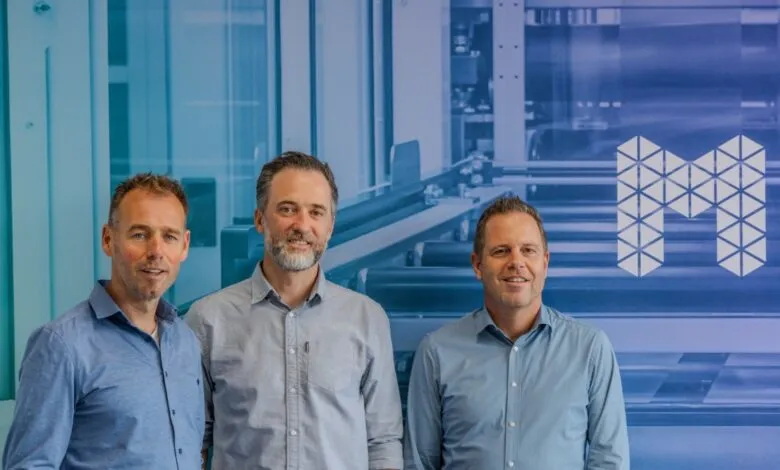 Three men in button-down shirts stand smiling in front of a blue industrial background.