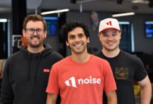 Three smiling men standing together inside a modern office, wearing casual clothing and branded apparel.