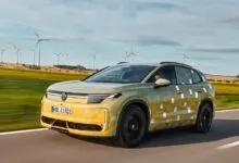 Camouflaged Volkswagen electric SUV driving on a country road past wind turbines.