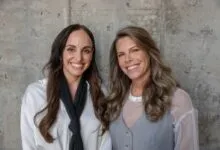 Two smiling women standing together against a textured concrete wall.