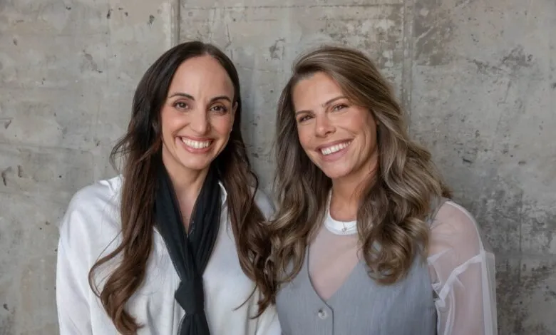 Two smiling women standing together against a textured concrete wall.