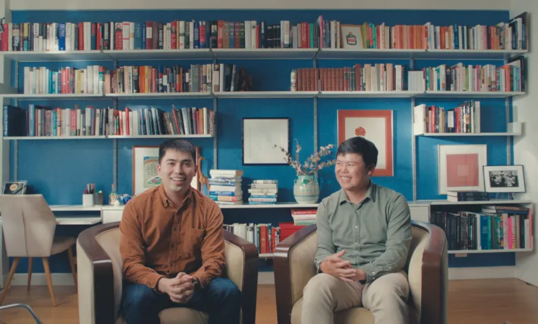 Two men seated in armchairs in front of a blue wall filled with loaded white bookshelves.