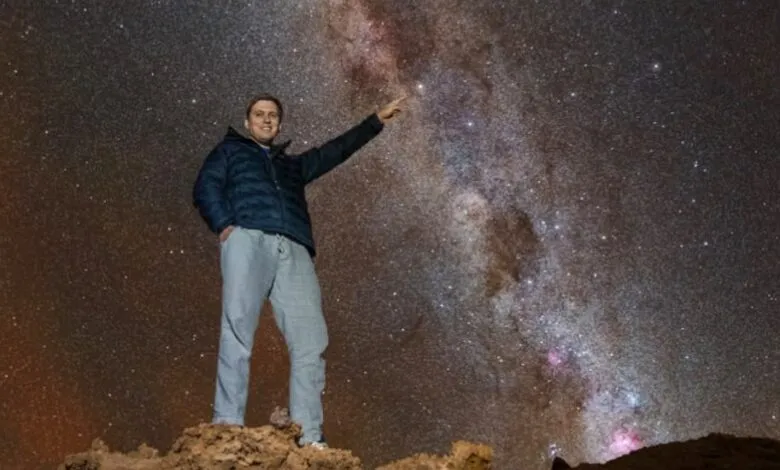 A man stands on a rocky outcrop at night, pointing up at the vast Milky Way galaxy stretching across the starry sky.
