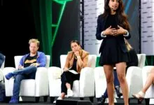 A woman speaks on stage during a TechCrunch Disrupt event while judges listen from white armchairs.