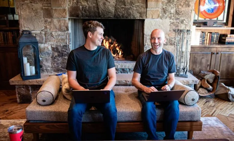 Two men laughing while working on laptops on a bench by a cozy stone fireplace.