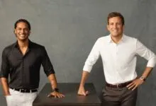 Two men standing confidently in front of a textured gray background, leaning on a dark table.