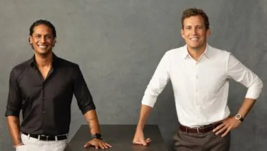 Two men standing confidently in front of a textured gray background, leaning on a dark table.