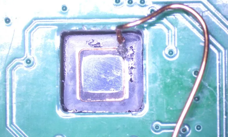 Microscopic view of a decapsulated RP2350 microcontroller chip on a green circuit board with attached wire.