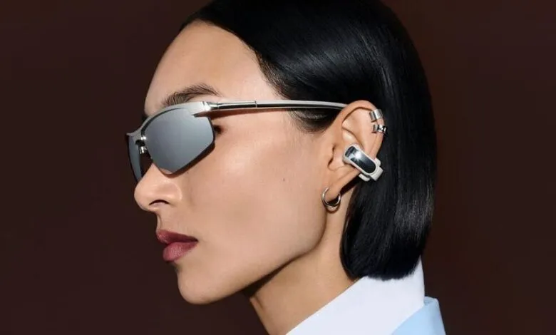 Close-up profile of a woman wearing silver sunglasses and innovative clip-on open-ear earbuds.