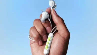A hand holds white Bose wired sports earbuds against a bright blue sky background.