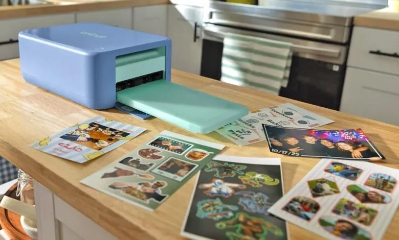 A light blue Cricut crafting machine on a wooden kitchen counter with printed stickers and photos spread around it.