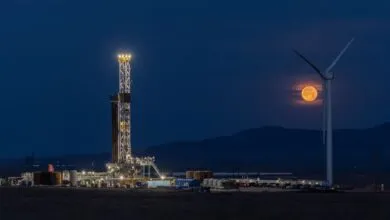 Fervo geothermal rig brightly illuminated at night beside a wind turbine and a large, glowing full moon.