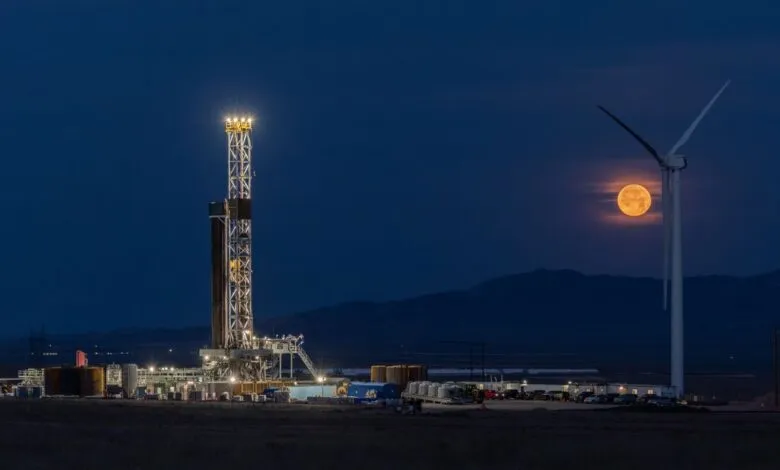 Fervo geothermal rig brightly illuminated at night beside a wind turbine and a large, glowing full moon.