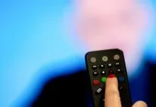 A finger with red nail polish presses a red record button on a black television remote control.