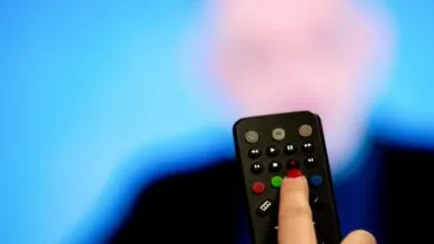 A finger with red nail polish presses a red record button on a black television remote control.