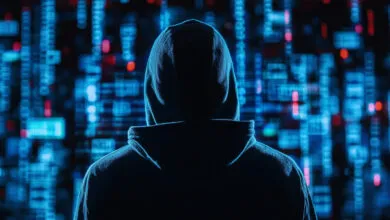 A hooded figure stands with their back turned against a glowing background of blue code.
