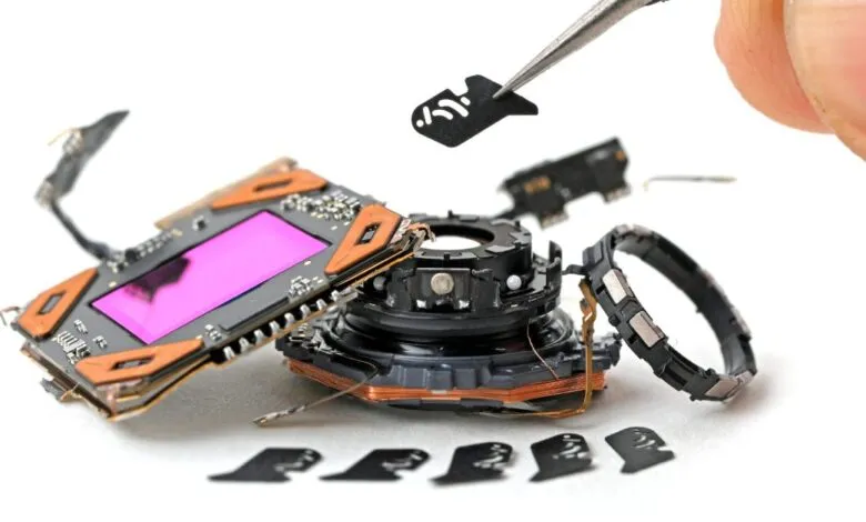 Tweezers carefully lift a small component from disassembled smartphone camera parts.