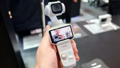 Hand holding a white Insta360 gimbal camera with an integrated screen showing a live view.