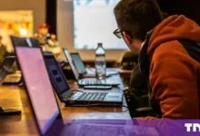Man wearing glasses and a red hoodie works on a laptop during a workshop with multiple computers.