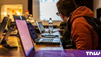 Man wearing glasses and a red hoodie works on a laptop during a workshop with multiple computers.