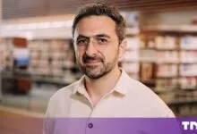 Portrait of Mustafa Suleyman wearing round glasses and a light-colored button-down shirt.