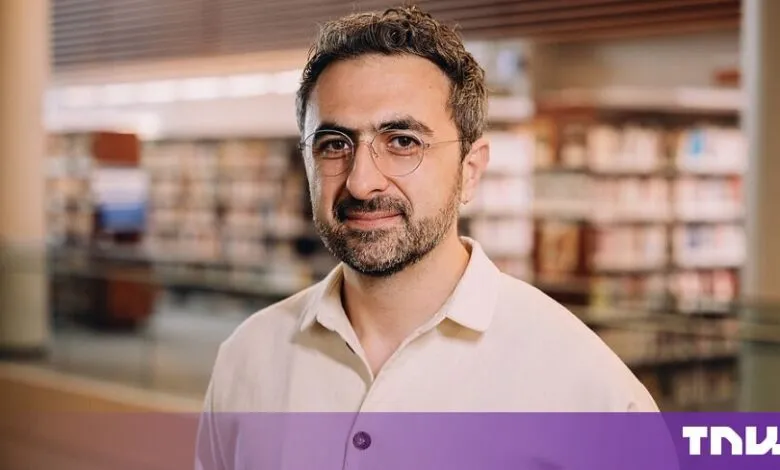 Portrait of Mustafa Suleyman wearing round glasses and a light-colored button-down shirt.