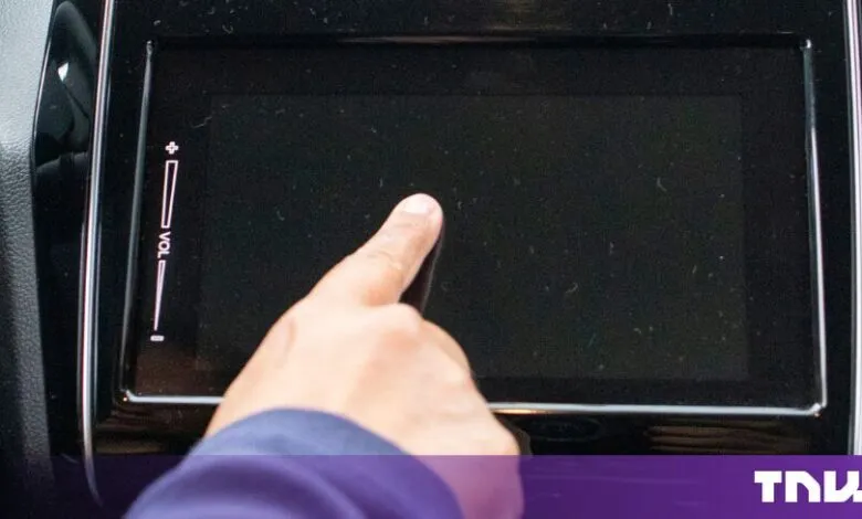 A finger touches a glossy, black car touchscreen display with a volume control slider